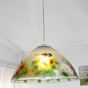 May include: A hanging pendant light with a frosted glass shade. The shade features a painted design of sunflowers, other flowers, and a small bird. The light fixture is suspended from a white ceiling.