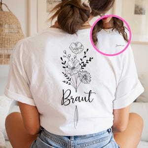 Op de afbeelding: Een wit t-shirt met een zwarte lijntekening van een bloemenboeket op de achterkant en het woord Braut onder de bloemen. Een cirkelvormige inzet toont de voorkant van het shirt met de naam Janina in zwarte cursieve letters.