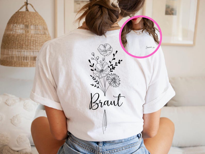 Op de afbeelding: Een wit t-shirt met een zwarte lijntekening van een bloemenboeket op de achterkant en het woord Braut onder de bloemen. Een cirkelvormige inzet toont de voorkant van het shirt met de naam Janina in zwarte cursieve letters.