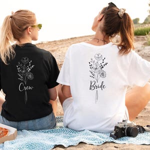 Two women sitting on a blanket on a beach, wearing matching t-shirts with floral designs and the text "Bride" printed on them.