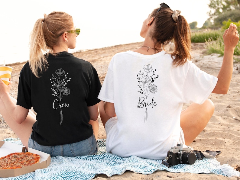 Two women sitting on a blanket on a beach, wearing matching t-shirts with floral designs and the text "Bride" printed on them.