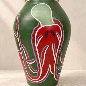 Könnte beinhalten: Eine grüne Keramikvase mit einem markanten roten Oktopus-Motiv, das mit hellblauen und weißen Linien umrandet ist. Die Vase hat einen bauchigen Körper und einen dicken, roten Rand an der Öffnung. Die Tentakel winden sich um die Seiten des Gefäßes.