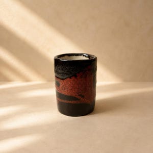 Può includere: Una tazza cilindrica in ceramica caratterizzata da uno smalto esterno nero scuro con una fascia testurizzata e maculata color bruno-rossastro che avvolge il centro. L'interno della tazza è rifinito con uno smalto liscio color bianco sporco.