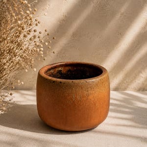 Op de afbeelding: Een cilindervormige keramische plantenpot met een gladde, ronde basis en een brede opening. De buitenkant heeft een verloop van warme, aardse tinten, variërend van diep, gebrand oranje aan de onderkant tot een lichtere, matte beige kleur.