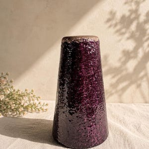 Op de afbeelding: Een conische keramische vaas met een dieppaarse, zwaar getextureerde en glanzende glazuurlaag. De bovenrand heeft een contrasterende lichtbruine, gespikkelde en matte afwerking. De vaas heeft een brede basis die naar boven toe smaller wordt.