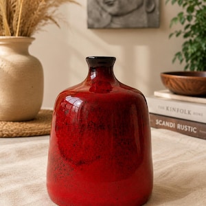 Può includere: Un vaso in ceramica rosso brillante e lucido con corpo arrotondato e collo stretto dal bordo scuro. La superficie presenta una finitura testurizzata e maculata con puntini rosso scuro e neri, creando un gradiente di colore ricco e profondo.