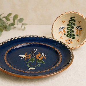 Op de afbeelding: Een donkerblauw keramieken bord en een crèmekleurige keramieken kom, beide met geschulpte randen en handgeschilderde bloemmotieven in oranje, groen en wit. Het bord heeft een centraal bloemmotief en de kom toont een verticaal groen patroon.