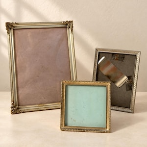 May include: Three rectangular and square picture frames with metallic gold and silver finishes. The largest frame has a dusty rose insert, the central square frame has a light blue insert, and the third frame shows a grey backing with a metal clip.