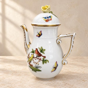 May include: A white porcelain teapot featuring a hand-painted design of colourful birds, green foliage, and butterflies. The lid is topped with a sculpted yellow rose finial. The handle and spout are decorated with gold trim and intricate patterns.