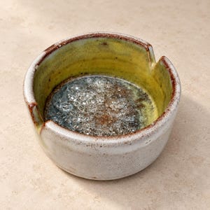 Pode incluir: Um cinzeiro redondo de cerâmica com exterior branco salpicado e interior com esmalte amarelo vibrante. A borda apresenta dois entalhes e um acabamento salpicado em castanho-avermelhado. A base contém fragmentos de vidro transparente e sedimentos escuros.