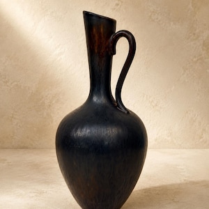 May include: A tall, dark brown ceramic pitcher with a bulbous body and a slender, elongated neck. The surface features a textured, mottled finish with subtle lighter brown streaks. It includes a single curved handle attached to the neck.