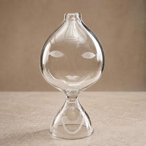 Könnte beinhalten: Eine Vase aus klarem Glas mit bauchigem Oberteil und ausgestelltem Fuß. Das Glas weist geätzte Details auf, darunter zwei mandelförmige Augen mit floralen Zentren, ein Paar Lippen und ein Perlenkettenmuster am unteren Sockelbereich.