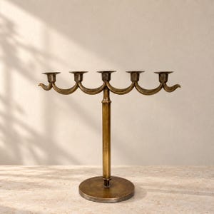 Könnte beinhalten: Ein Messing-Kandelaber mit einem zentralen vertikalen Stiel, der auf einem runden Sockel ruht. Der horizontale Arm trägt fünf Kerzenhalter, die in einem symmetrischen, geschwungenen Design mit spitz zulaufenden Enden angeordnet sind.