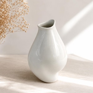 Peut inclure: Un vase en céramique blanche lisse avec une forme organique, asymétrique et bulbeuse. Le récipient présente une ouverture étroite et légèrement irrégulière au sommet ainsi qu'un corps arrondi et incurvé qui s'affine vers la base.