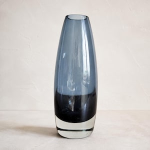 Può includere: Un vaso in vetro alto e affusolato con un design a gradiente di colore. La sezione superiore è grigio chiaro traslucido, che sfuma in una profonda fascia nera opaca al centro, terminando con una base in vetro spesso e trasparente.
