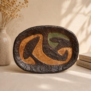 Può includere: Un vassoio ovale in ceramica con un bordo esterno marrone scuro, fortemente strutturato e ruvido. Il centro presenta un design astratto con zone lisce e contrastanti di colore beige chiaro e verde salvia su uno sfondo marrone scuro.
