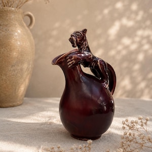 Può includere: Una brocca in ceramica color bordeaux scuro con una figura felina scolpita appollaiata sul manico. La brocca presenta una finitura liscia e lucida, un corpo arrotondato e un beccuccio svasato. La figura felina è integrata nel manico.