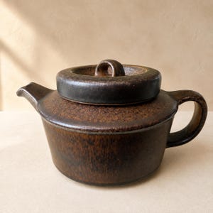 Könnte beinhalten: Eine dunkelbraune Keramik-Teekanne mit einer gesprenkelten, strukturierten Glasuroberfläche. Die Teekanne verfügt über einen breiten, flachen Deckel mit einem zentralen Griff, einen kurzen Ausguss links und einen geschwungenen Henkel rechts.