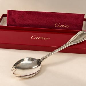 Könnte beinhalten: Ein silberfarbener Servierlöffel aus Metall mit einer eingekerbten Laffe und einem gerippten Griff, präsentiert in einer roten Cartier-Box. Die Box hat einen glänzenden roten Deckel mit goldenem Cartier-Logo und dunkelrotem Samtfutter.