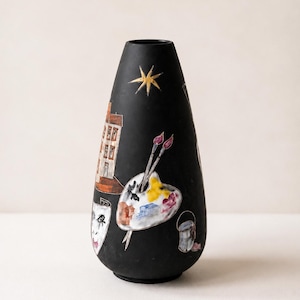 Pode incluir: Um vaso preto fosco de formato cônico com ilustrações pintadas à mão, incluindo uma estrela dourada, um edifício, uma paleta de artista branca com manchas de tinta coloridas e dois pincéis, uma máscara de teatro e um pequeno balde de tinta.