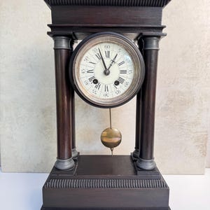 Könnte beinhalten: Eine dunkelbraune Holzkaminuhr mit einem runden Zifferblatt, schwarzen römischen Ziffern und arabischen Minutenmarkierungen. Die Uhr wird von vier Säulen getragen und verfügt über ein zentrales Messingpendel unter dem Zifferblatt auf einem rechteckigen Sockel.