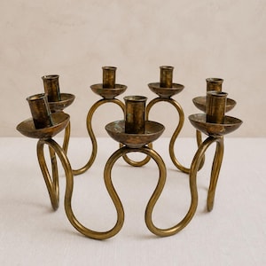 May include: A circular brass candelabra featuring six individual candle holders connected by a continuous, undulating, wave-like metal frame. The brass has a weathered, aged patina with visible dark spots and a dull, metallic finish throughout.