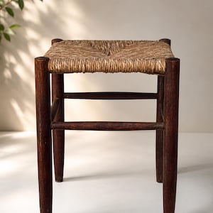 Könnte beinhalten: Ein quadratischer Hocker mit vier dunkelbraunen Holzbeinen, die durch horizontale Stützstreben verbunden sind. Die Sitzfläche besteht aus geflochtenen, naturfarbenen Fasern, die eine strukturierte Oberfläche bilden. Der Hocker ist schlicht.