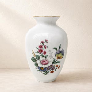 Könnte beinhalten: Eine weiße Porzellanvase mit goldfarbenem Rand, die mit einer zentralen botanischen Illustration verschiedener Blumen verziert ist, darunter rosa, weiße, blaue und violette Blüten mit grünen Blättern, die in einem Bündel auf dem bauchigen Körper angeordnet sind.