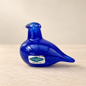 May include: A vibrant cobalt blue glass bird figurine with a smooth, rounded body and a pointed tail. A silver and blue sticker on the side features the text NUUTAJÄRVI 1793 MADE IN FINLAND. The glass has a glossy finish and a clear, defined beak.