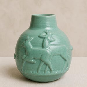 Peut inclure: Un vase en céramique vert clair de forme ronde, orné d'un relief représentant deux cerfs dans une prairie. Le vase présente une finition émaillée lisse avec une texture craquelée subtile et une ouverture circulaire étroite au sommet.