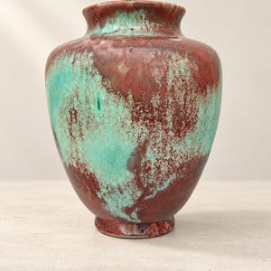 Könnte beinhalten: Eine Keramikvase mit bauchigem Körper und ausgestelltem Rand, die eine gesprenkelte Glasur aufweist, welche tiefe rotbraune Töne mit leuchtend türkisfarbenen Flecken vermischt und so ein strukturiertes, geschichtetes Erscheinungsbild erzeugt.