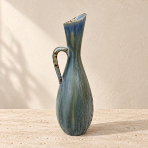 Può includere: Un alto vaso a brocca in ceramica con corpo a forma di goccia e collo sottile e svasato. La superficie presenta uno smalto sfumato nei toni del blu profondo, verde salvia e marrone terroso con un motivo a goccia verticale e un manico curvo.