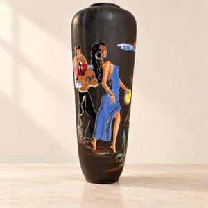 Può includere: Un alto vaso cilindrico nero opaco caratterizzato da una scena dipinta a colori che raffigura una persona con un abito blu che tiene un tamburello e un'altra con una camicia rossa che suona la chitarra. L'opera presenta vivaci accenti blu, rossi e gialli.