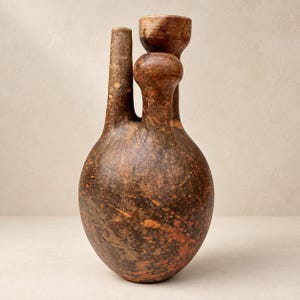 Pode incluir: Um recipiente de cerâmica castanho e manchado com uma base arredondada e dois bicos verticais distintos de alturas diferentes. A superfície apresenta um acabamento texturizado e desgastado com variações de cor castanho escuro e laranja avermelhado.
