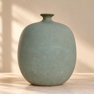 Pode incluir: Um vaso redondo de cor verde sálvia com um acabamento mate suave e um pescoço estreito. A superfície do vaso apresenta imperfeições naturais subtis e pequenos poros, conferindo-lhe um aspeto texturado mantendo uma forma arredondada.