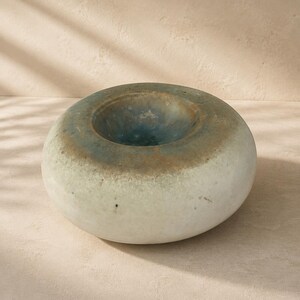May include: A low, rounded ceramic vessel with a smooth, off-white base that transitions into a mottled, earthy brown and deep blue-green glaze around the central circular opening, creating a textured, multi-toned appearance on the surface.