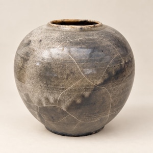 Może przedstawiać: Okrągłe ceramiczne naczynie z szerokim korpusem i wąskim otworem. Powierzchnia posiada nakrapianą szaro-brązową glazurę z wyraźną siecią cienkich białych linii spękań na całej zewnętrznej stronie, co nadaje jej teksturowany wygląd.