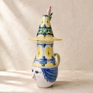 Può includere: Una brocca in ceramica con un volto dipinto a mano, capelli blu, naso a punta e una piccola bocca rossa. La brocca è sormontata da un coperchio conico decorato con motivi geometrici gialli, verdi e blu, rifinito con un piccolo pomello rosa.