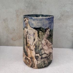 Pode incluir: Um vaso cilíndrico de cerâmica com uma ilustração pintada à mão de duas figuras numa paisagem. A obra utiliza tons suaves de azul, verde e bege com contornos escuros. A borda interior é azul-escura com um padrão circular branco.