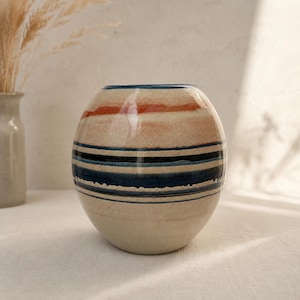 Pode incluir: Um vaso de cerâmica redondo com uma base lisa em bege claro. O exterior apresenta faixas horizontais de cor, incluindo uma risca laranja fina perto do topo e uma série de faixas mais grossas e texturizadas em azul escuro e preto no centro.