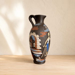 Pode incluir: Um jarro de cerâmica castanho-escuro com uma cena pintada à mão de uma pessoa sentada num banco perto de uma parede de tijolo e de uma porta de pedra em arco. O design inclui detalhes em azul, branco e laranja, com uma pega lateral.