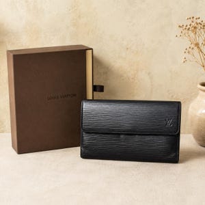 Może przedstawiać: Czarny portfel Louis Vuitton z teksturowanym wzorem poziomych linii i małym wytłoczonym logo LV w prawym przednim rogu, któremu towarzyszy pasujące brązowe prostokątne pudełko do przechowywania z nadrukowaną nazwą marki Louis Vuitton.