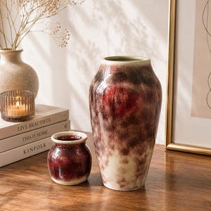 Könnte beinhalten: Eine hohe, konische Keramikvase und ein kleinerer, bauchiger Keramiktopf, beide mit einer gesprenkelten, reaktiven Glasur in den Farben Dunkelrot, Dunkelbraun und Creme. Die Oberflächen haben ein glänzendes Finish mit ungleichmäßigen Übergängen.