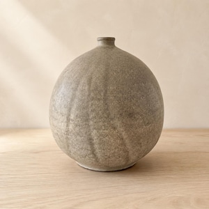 Può includere: Un vaso in ceramica sferico con una finitura opaca e testurizzata color tortora. La superficie presenta sottili scanalature verticali e un'apertura circolare stretta sulla parte superiore. L'oggetto ha un corpo rotondo e una base piatta.