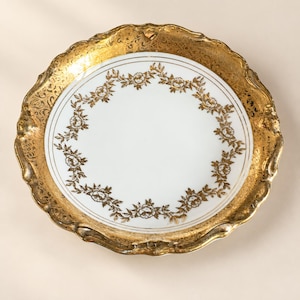Op de afbeelding: Een ronde porseleinen bord met een wit middenstuk versierd met een cirkelvormige gouden bloemenslinger. De brede, geschulpte rand is afgewerkt met een sierlijk, getextureerd goudpatroon, wat een duidelijk contrast vormt met het wit.