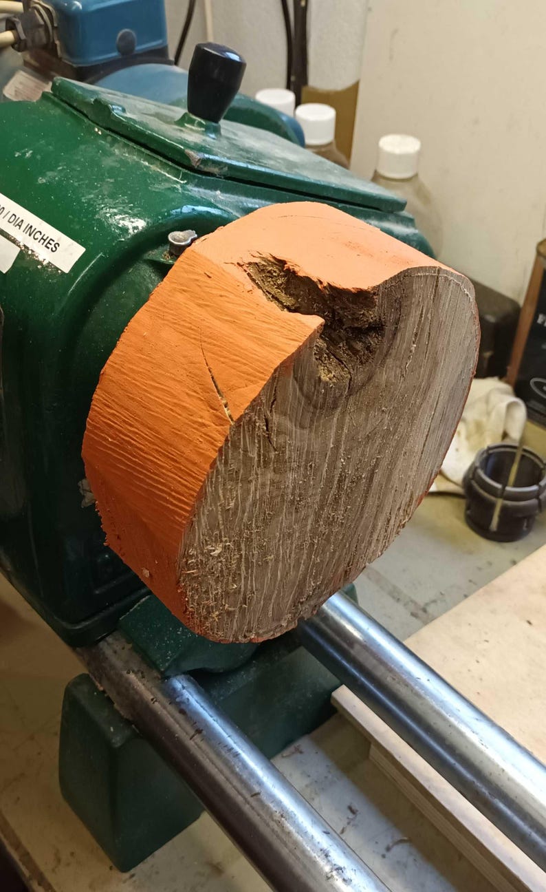 Może przedstawiać: Zbliżenie na tokarkę do drewna z zamontowanym na niej kawałkiem drewna. Drewno jest pomalowane na pomarańczowo i ma sęk. Tokarka jest zielona i ma metalowy uchwyt.