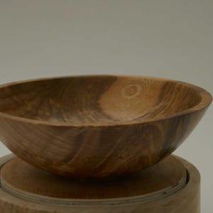Może przedstawiać: Duża drewniana miska z naturalnym brązowym wykończeniem. Miska ma szeroką, zaokrągloną podstawę i głębokie, zakrzywione wnętrze. Widoczne usłojenie drewna dodaje misce rustykalnego uroku.