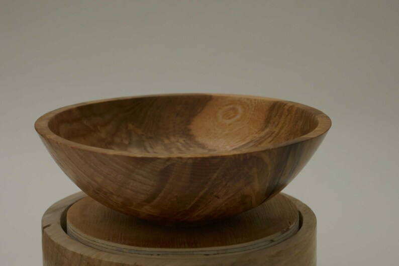 Może przedstawiać: Duża drewniana miska z naturalnym brązowym wykończeniem. Miska ma szeroką, zaokrągloną podstawę i głębokie, zakrzywione wnętrze. Widoczne usłojenie drewna dodaje misce rustykalnego uroku.