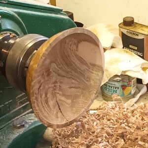 Może przedstawiać: Drewniana miska obracana na tokarce. Miska jest jasnobrązowa z ciemnobrązowymi smugami. Tokarka jest zielona i ma czarny metalowy uchwyt. Na podłodze znajdują się wi&oacute;ry drzewne.