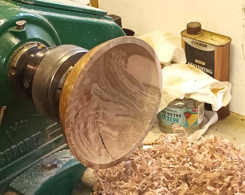 Może przedstawiać: Drewniana miska obracana na tokarce. Miska jest jasnobrązowa z ciemnobrązowymi smugami. Tokarka jest zielona i ma czarny metalowy uchwyt. Na podłodze znajdują się wi&oacute;ry drzewne.